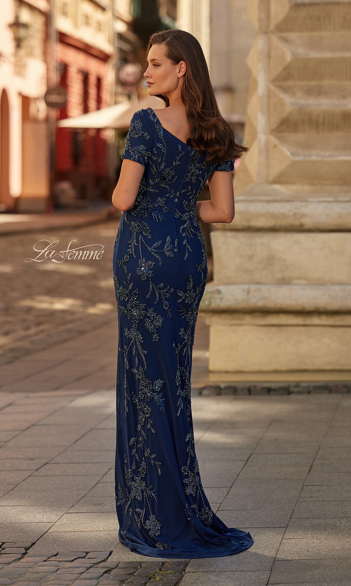 A woman stands outdoors on a cobblestone street, wearing the Long Formal Dress: La Femme 33577—a fitted navy gown with beaded floral embroidery and short sleeves—her back elegantly turned to the camera.